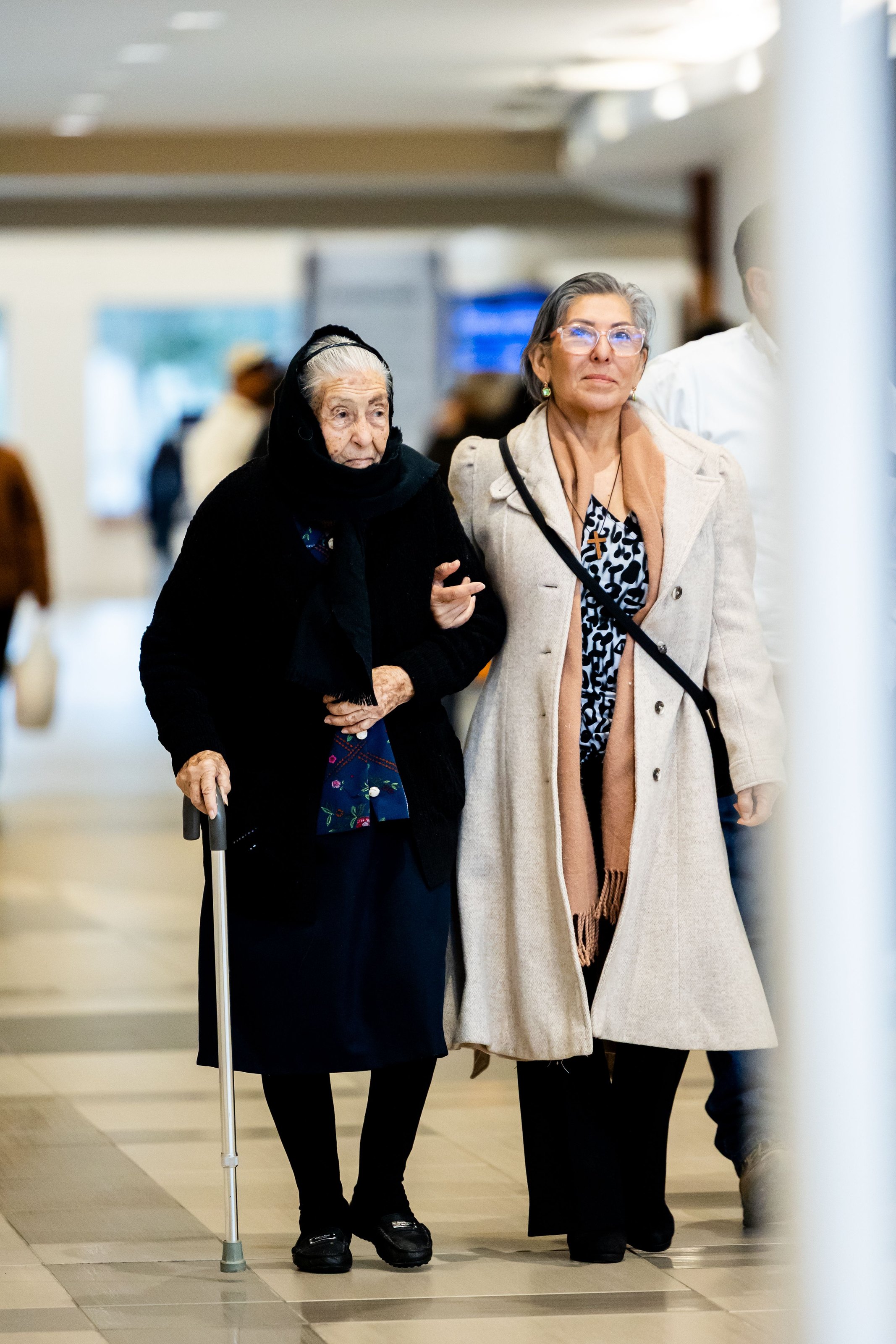 An elderly woman with a walking stick, arm in arm with a younger woman, walking through a terminal