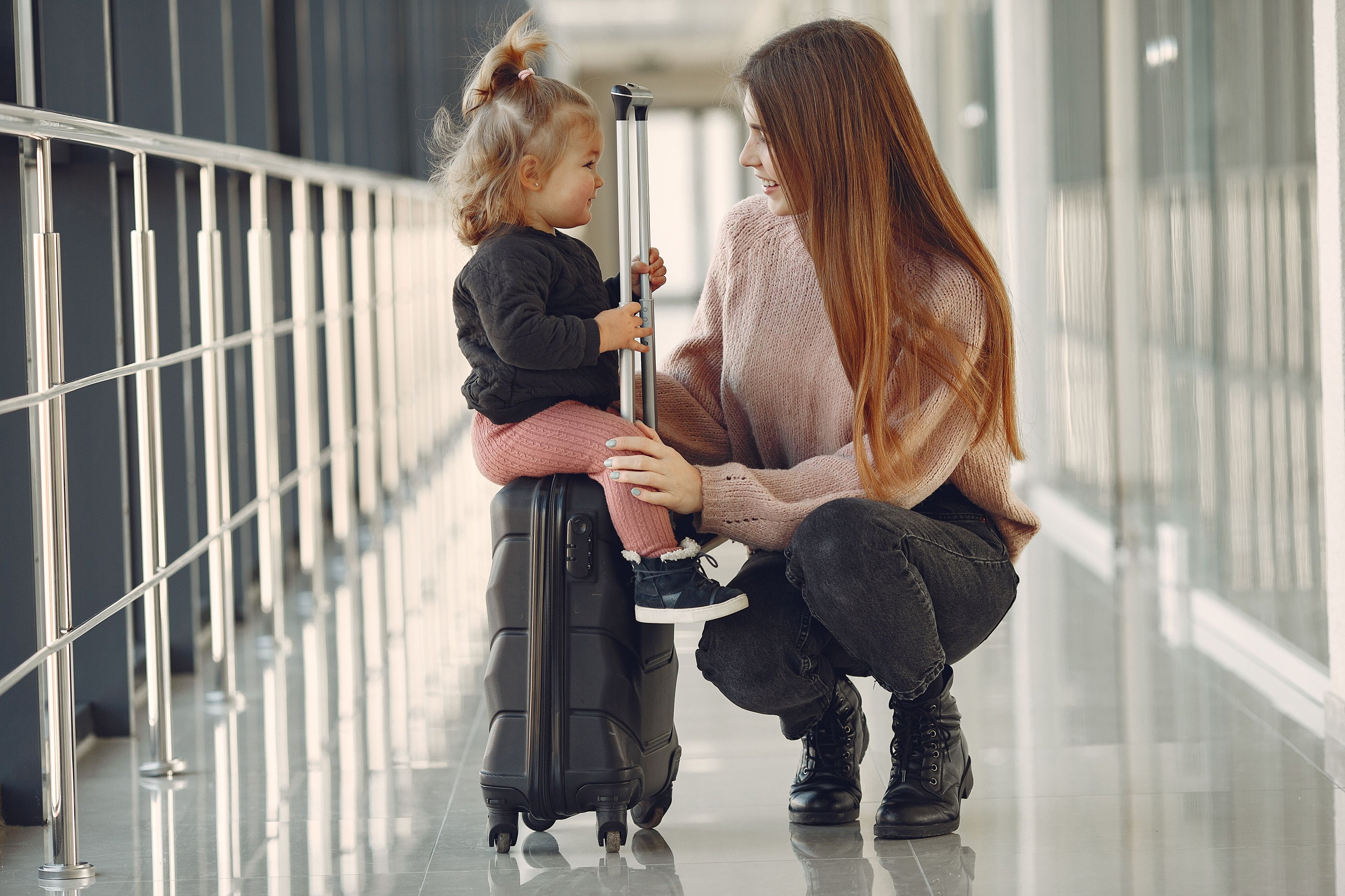 A woman kneeling beside a little girl sitting on a suitcase in an airport corridor