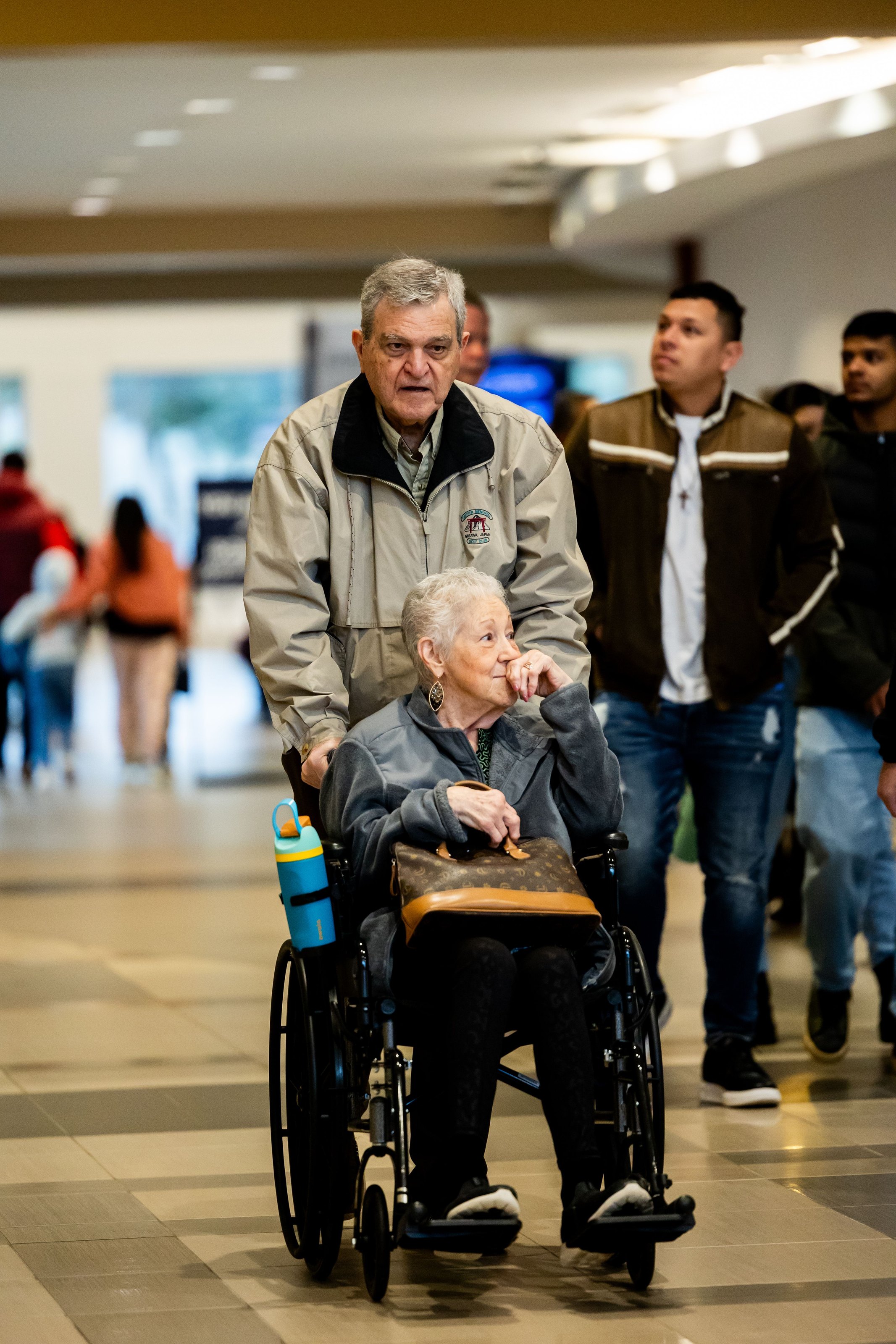 An elderly woman in a wheelchair being helped through a busy shopping centre
